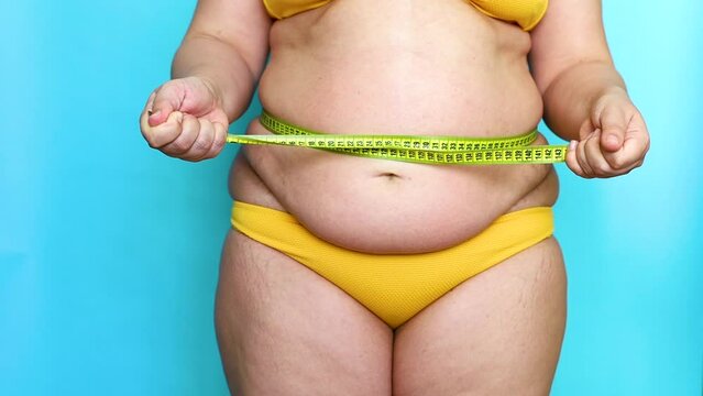 Unrecognizable plump overweight woman standing in yellow bra, swimming trunks, measuring waist with tape, tightening, breathing in to reduce size. Body positive, cellulite, obesity, weight control.