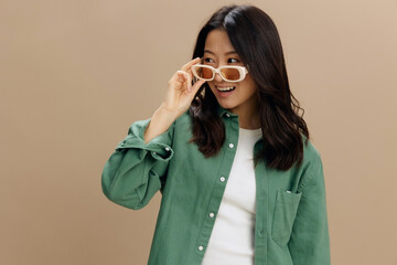 Happy enjoyed Korean young woman in khaki green shirt stylish eyewear looks aside posing isolated on over beige pastel studio background. Cool fashion offer. Sunglasses ad concept
