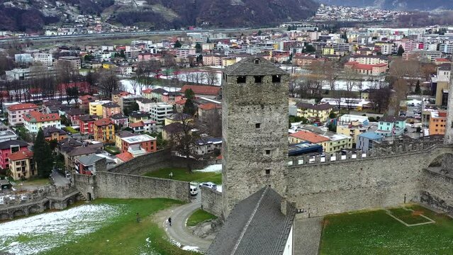 Aerial drone footage of a medieval castle in Europe.  High castle walls and towers, with the village below.  Surrounded by snowy mountains with an overcast sky. 