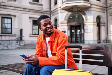 A young man sitting on a bench in the city with his luggage and holding his phone.