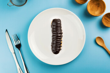 Dessert Bomba de Chocolate. Brazilian traditional éclair in blue plate on the yellow table. Top view. Kitchen utensils beside like fork, knife, bamboo containers
