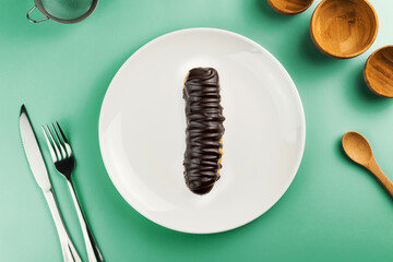 Dessert Bomba de Chocolate. Brazilian traditional éclair in green plate on the yellow table. Top view. Kitchen utensils beside like fork, knife, bamboo containers