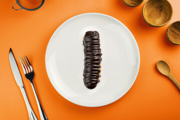 Dessert Bomba de Chocolate. Brazilian traditional éclair in orange plate on the yellow table. Top view. Kitchen utensils beside like fork, knife, bamboo containers.