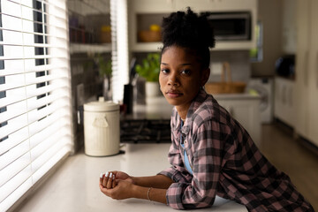 Portrait of serious african american young woman leaning on kitchen counter while standing at home