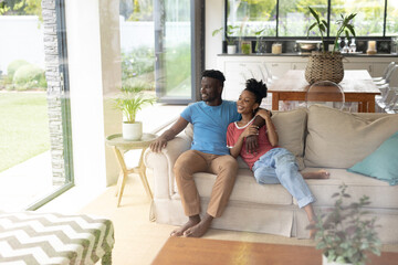 Smiling african american young couple looking through window while relaxing on sofa in living room