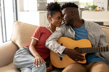 Loving african american young man looking at girlfriend while playing guitar on sofa in living room