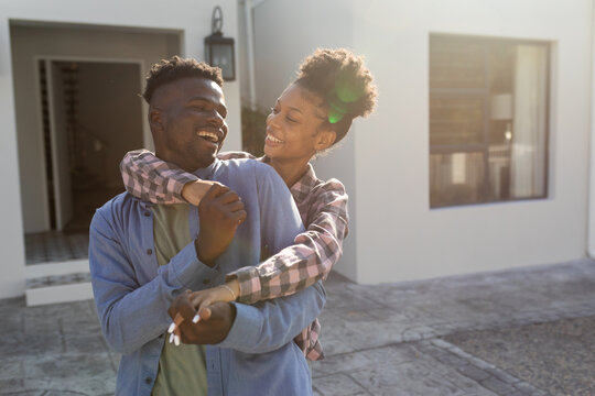 Loving african american young woman embracing and holding cheerful boyfriend's hands against house