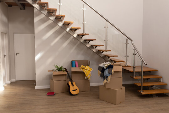 Stack of cardboard boxes with guitar, books and clothing by staircase in new empty house, copy space