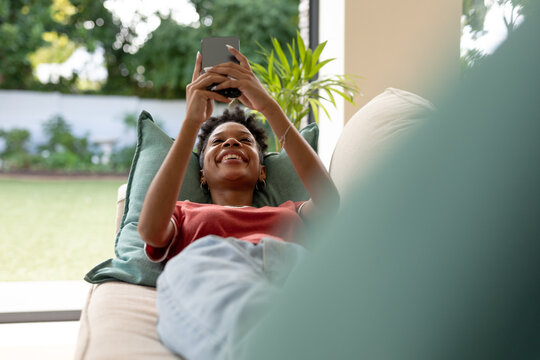 Smiling african american young woman using mobile phone while lying on sofa in living room at home