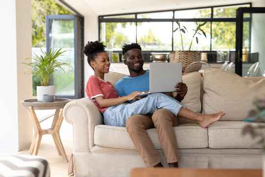 Cheerful african american young couple using laptop while sitting on sofa in living room at home