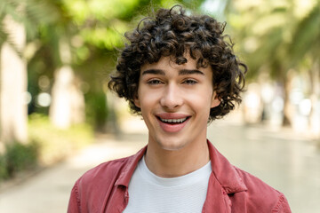 Portrait of cheerful young curly man standing in city park on sunny warm day and smiling