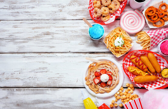 Carnival theme food side border over a white wood background. Above view with copy space. Summer fair concept. Corn dogs, funnel cake, cotton candy and snacks.