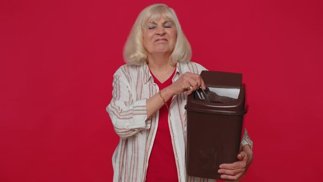 Happy senior woman 70s in shirt taking off, throwing out glasses into bin after vision laser treatment therapy, looking smiling at camera. Elderly grandmother isolated alone on red studio background