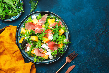 Delicious summer fresh salad with cantaloupe melon, prosciutto, soft cheese and arugula on blue table background, top view, copy space