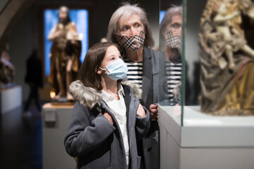 Fototapeta na wymiar Friendly elderly female tutor in protective face mask showing interested tween girl exposition of artworks in museum of archeology and ancient sculptures. New normal in coronavirus pandemic
