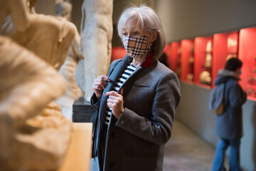 Portrait of interested aged woman wearing protective face mask examining ancient sculptures while...