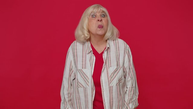 Cheerful funny senior woman in shirt showing tongue making faces at camera, fooling around, joking, aping with silly face, teasing. Elderly old grandmother isolated alone on red studio wall background
