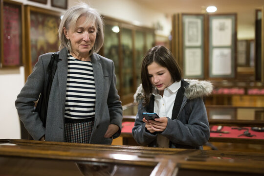 Cute curious tween girl browsing internet on smartphone in search of information about showpieces while visiting local history museum with her grandmother