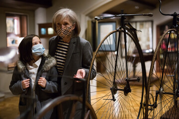 Curious tween schoolgirl and friendly elderly female tutor in face masks viewing vintage bicycle in...