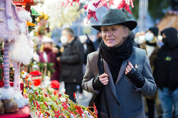 Positive stylish elderly woman in festive mood enjoying walking on street Christmas fair, choosing decorations for home