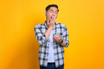 Bored young Asian man in plaid shirt holding mobile phone and feeling lack of sleep isolated on yellow background