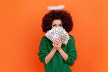 Portrait of angelic woman with Afro hairstyle wearing green casual style sweater and nimb covering half of face with big fan of dollars banknotes. Indoor studio shot isolated on orange background.