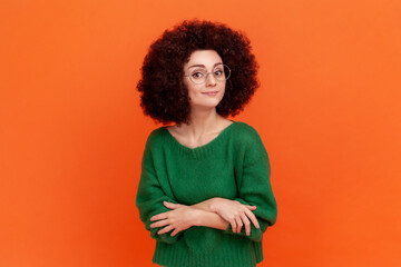 Portrait of confident woman with Afro hairstyle wearing green casual style sweater and optical glasses, standing with folded arms, looking at camera. Indoor studio shot isolated on orange background.