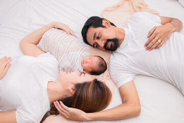 Obraz na płótnie Canvas Asian young mother father and newborn lying on bed together. Mom and dad happy parents sleep next to their little baby. Little infant deeply sleeping wrapped in thin white cloth with happy and safe.