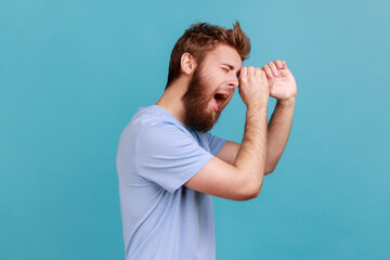 Side view of wondered shocked bearded man making monocular gesture with hands and looking amazed at side space, keeps mouth opened. Indoor studio shot isolated on blue background.