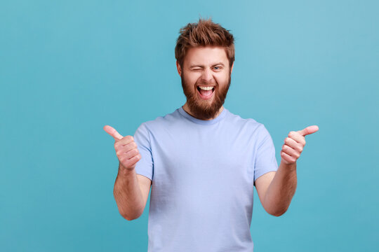 Portrait of bearded man approves incredible promo keeps thumbs up likes and agrees being satisfied with something, winking to camera. Indoor studio shot isolated on blue background.