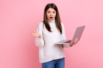 Portrait of attractive shocked girl standing with laptop in hands, looking at camera with open mouth, wearing white casual style sweater. Indoor studio shot isolated on pink background.