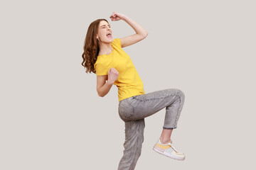 Full length excited teenager girl in casual yellow T-shirt and grey jeans posing, clenching fists...
