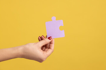Profile side view closeup of woman hand holding one purple puzzle piece, solving tasks. Indoor studio shot isolated on yellow background.