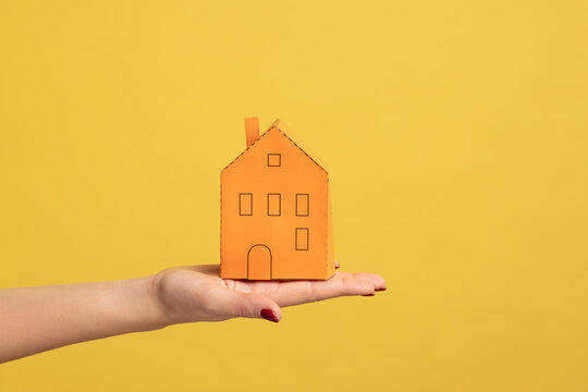 Real estate insurance, repairing services. Closeup of woman hand holding small orange paper house, concept of dream home purchase, mortgage. Indoor studio shot isolated on yellow background.