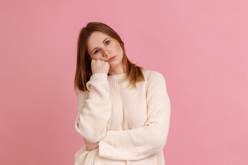 Portrait of blond woman leaning on hand and expressing disinterest apathy with dull look, feeling lazy indifferent, wearing white sweater. Indoor studio shot isolated on pink background.