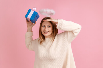 Portrait of happy blond woman with nimb over head holding gift box in hands and looking far, finding who gives a present, wearing white sweater. Indoor studio shot isolated on pink background.