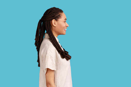 Side view portrait of cheerful woman with black dreadlocks, having positive optimistic emotions and charming engaging smile, wearing white shirt. Indoor studio shot isolated on blue background.