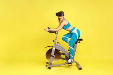 Fototapeta na wymiar Side view portrait of athletic sporty woman pedaling on exercise bike looking through augmented reality viewer, wearing blue sportswear. Indoor studio shot isolated on yellow background.