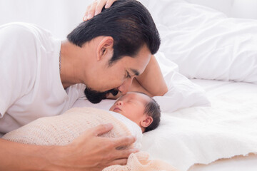 Fototapeta na wymiar Close up of Asian young father kissing newborn while baby deeply sleeping with happiness. Little infant wrapped in thin white cloth with happy and safe. Dad and toddler spend time together.