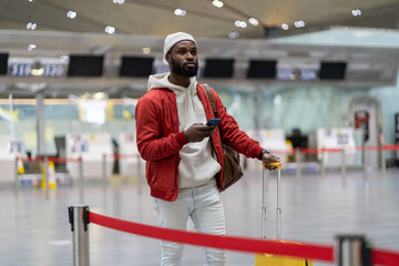 Thoughtful African American passenger man waiting to check in and drop off luggage in airport terminal, checks time of flight online, stands with suitcase, using mobile phone. Tourism, traveling. 