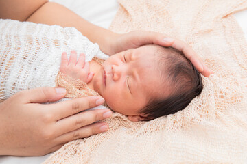 Asian little newborn baby deeply sleeping wrapped in thin white clothwith happy and safe on beige blanket with touching of mother's care. Mom touch adorable infant with love and tender.