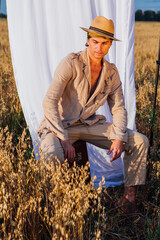 Tall handsome man dressed in a coarse linen suit and hat sitting on the old vintage suitcase at golden oat field with a white curtain on the background.