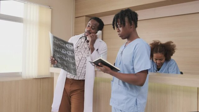 Professional African-American male doctor talks phone about a patient's diagnosis X-ray film and medicine at reception counter of hospital's outpatient clinic, and young medical students together.