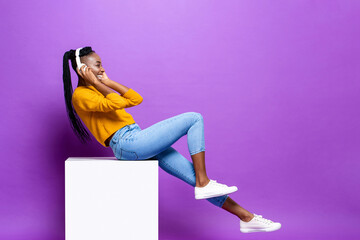 Smiling happy young African-American woman wearing headphone sitting on stool listening to music in...