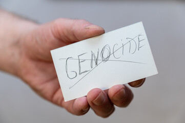 The word GENOCIDE is crossed out. A word written in jagged letters. A man's hand holds a white paper rectangle with text against a gray background
