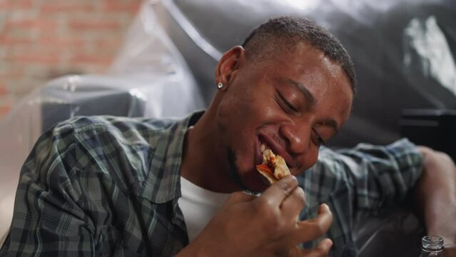 Smiling Afro-American guy bites pizza slice leaning on sofa