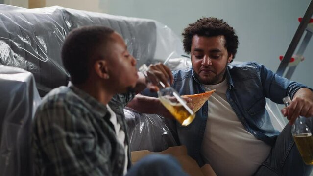 Positive Afro-American guys enjoy beer and pizza near sofa