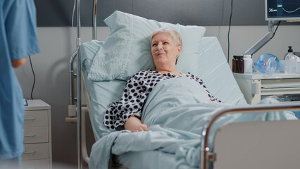 Medical assistant doing checkup visit for aged patient in hospital ward bed. Woman working as nurse...