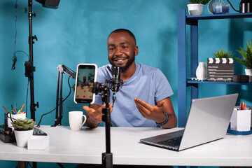 Smiling vlogger talking with audience in front of recording smartphone during online live show...