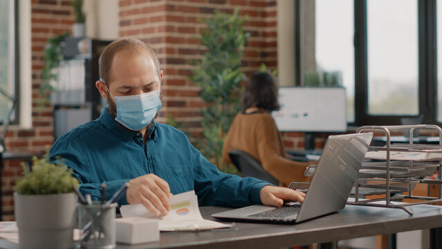 Employee wearing face mask and analyzing charts papers while working with laptop on business project. Entrepreneur using technology and files to work on presentation in startup office.